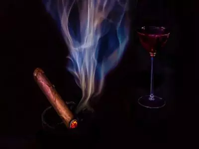 A lit cigar with lots of smoke and a wine glass in the background