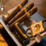 Couple of cigars sitting in a tray with a cigar cutter.